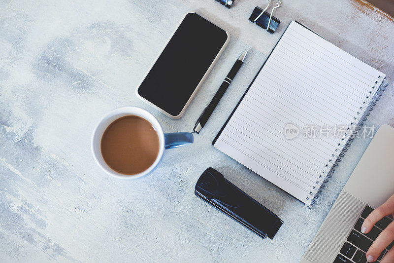 Top view of woman using laptop while sitting at the table with laptop, mobile phone, blank note pad and pen.女人使用笔记本电脑时的俯视图。